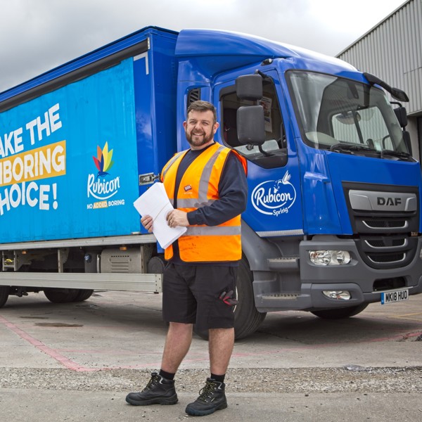 A Barr Soft Drinks delivery driver stands smiling in front go a branded Rubicon lorry